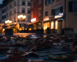 Before the Lights Fade A tranquil dawn street photograph capturing fallen autumn leaves on cobblestones beneath the soft glow of early café lights. Warm tones meet the cool morning...