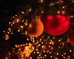 Ember of Winter Warm close-up of red and gold Christmas ornaments hanging from pine branches, illuminated by soft golden fairy lights with rich bokeh in the background.