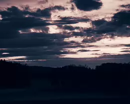 Fading Quietude A moody twilight landscape captured near Frauenfeld, Switzerland, featuring dramatic layered clouds and a subtle evening glow above a silhouetted treeline. The...