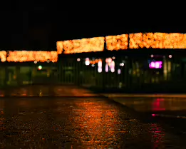Glowline Low-angle night photograph showing warm orange holiday lights blurred into a horizontal ribbon above a wet pavement, with soft neon reflections in pink and...