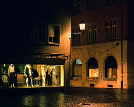 The Still Hour A quiet town square after dark, illuminated by a shop window and a solitary street lamp. Rain-soaked cobblestones reflect the warm glow, turning an empty scene...