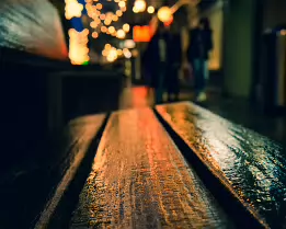 Where the Night Rests Rain-darkened wood catches the glow of distant streetlights, turning a simple bench into a quiet stage for the city’s passing life. Soft bokeh and silhouettes...