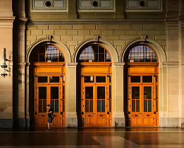 Warm Portals A lone man walks past ornate wooden double doors framed by stone arches, illuminated by warm dawn light.