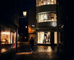 Crossing the Quiet Glow A moody night-street photograph capturing a solitary figure walking past softly illuminated storefronts and cobblestone reflections. The warm streetlights and...