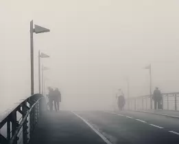 Fog Crossing Pedestrians and a cyclist cross a fog-shrouded bridge lined with evenly spaced lamps.